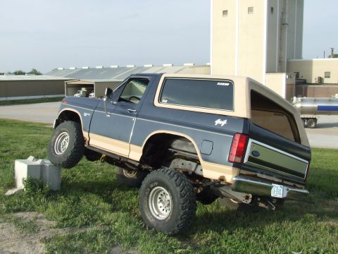1986 Ford Bronco - Lifted two-tone blue and tan full-size Ford Bronco flexing its suspension with the driver-front tire up on a concrete block, showing articu…