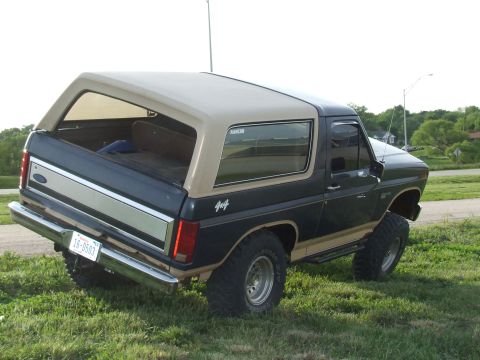1986 Ford Bronco - Driver-side rear three-quarter view of a lifted two-tone dark blue and tan full-size Ford Bronco on grass, with the tailgate glass retracte…