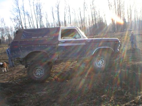 1978 Ford Bronco - A 1978 Ford Bronco is seen from the side, navigating a muddy, wooded terrain under bright sunlight.