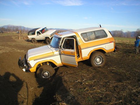 1978 Ford Bronco - A white and orange 1978 Ford Bronco is stuck in the mud, its driver's side door ajar, while another off-road vehicle is visible in the back…