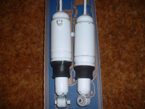 1978 Ford Mustang II - Two white shock absorbers are displayed against a textured brown background and a blue box with striped detailing.