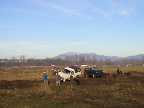 1978 Ford Bronco - Multiple off-road vehicles are bogged down in a muddy terrain under a clear sky with mountains in the distance.