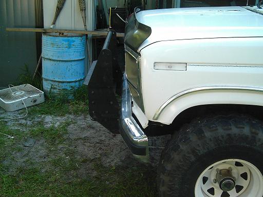 White Ford truck front end with chrome bumper and large tires. Blue barrel and fan in background
