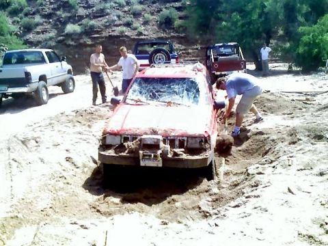 1991 Ford Bronco - Several people are attempting to free a red vehicle that is stuck in a deep rut of dirt.