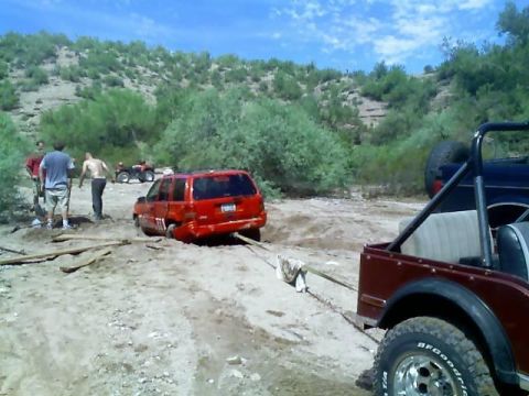 1991 Ford Bronco - Red SUV stuck in mud, being pulled by a red Jeep. People and ATVs in background.