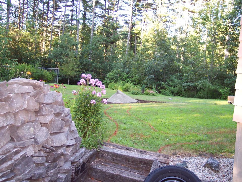 Backyard scene with a stacked stone wall in the foreground, pink flowers, a pile of sand, and trees in the background