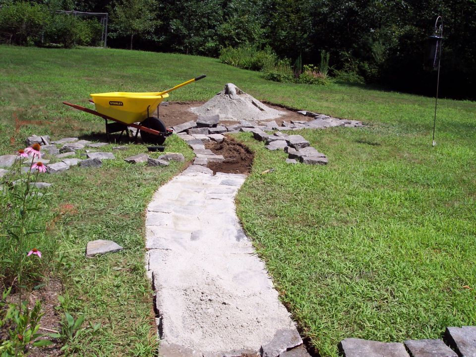 Yellow wheelbarrow next to pile of sand and scattered stones for garden path construction