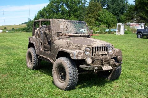 1978 Ford Bronco - Mud-covered Jeep Wrangler parked on a grassy field with trees and a small building in the background.