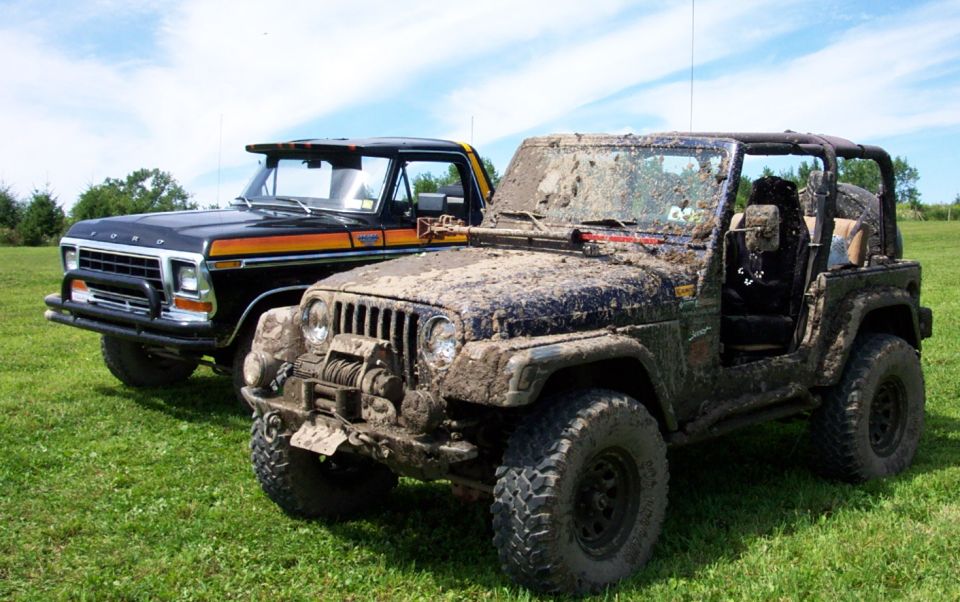 Muddy Jeep Wrangler parked next to a black Ford pickup with orange stripes. Both vehicles are on a grassy field