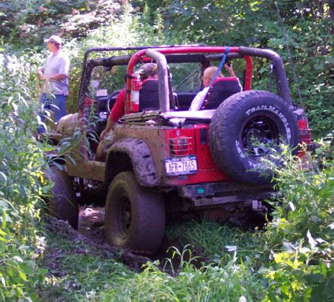 1978 Ford Bronco - A red Jeep Wrangler navigates a muddy, overgrown trail with people inside.