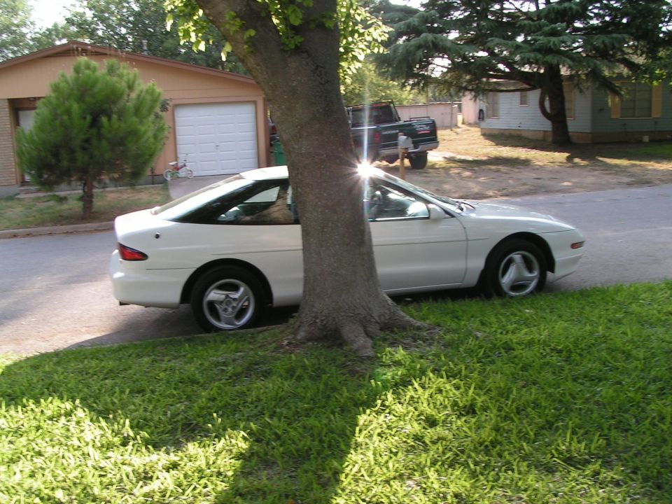 White Ford Probe coupe in side view parked at a curb behind a large tree, with houses in the background