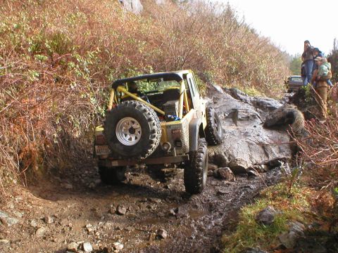 1978 Ford Bronco - A yellow off-road vehicle tackles a challenging muddy trail with other vehicles and people in the background.