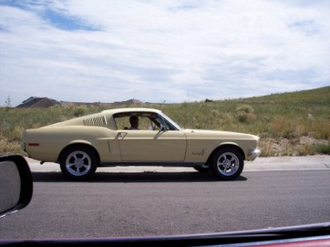 1972 Ford Ranchero - A pale yellow 1967 Ford Mustang Fastback is seen from the side, driving on a road with a grassy hill in the background.
