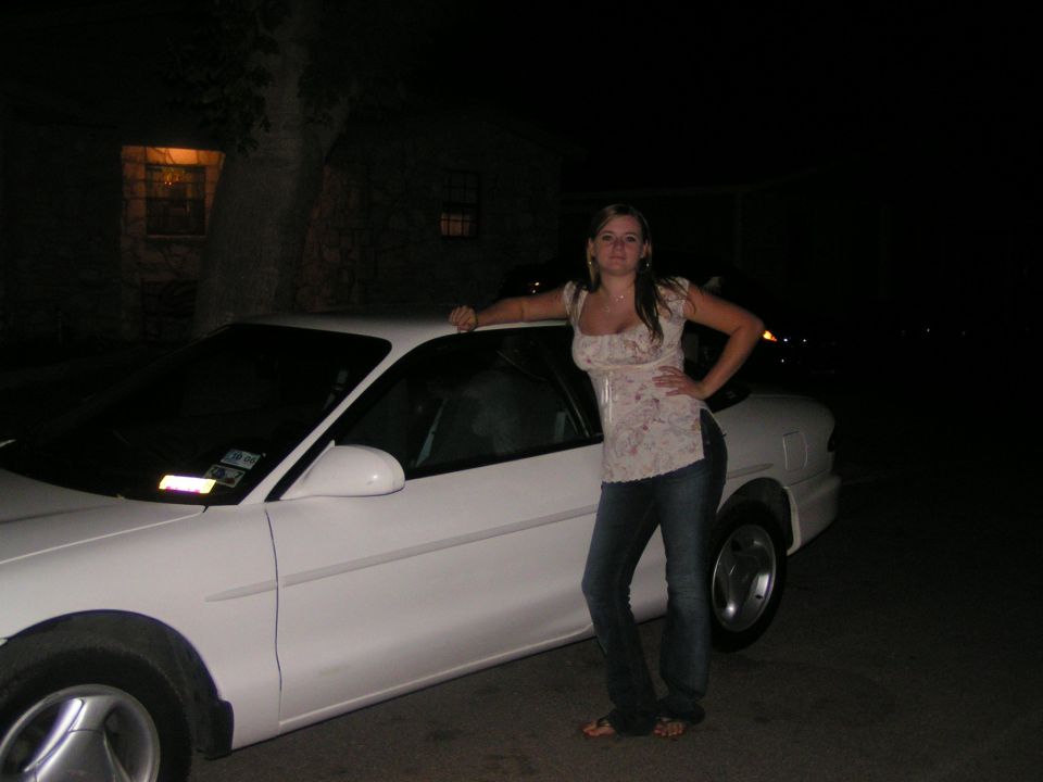Woman posing beside a white coupe at night, arm resting on the roof