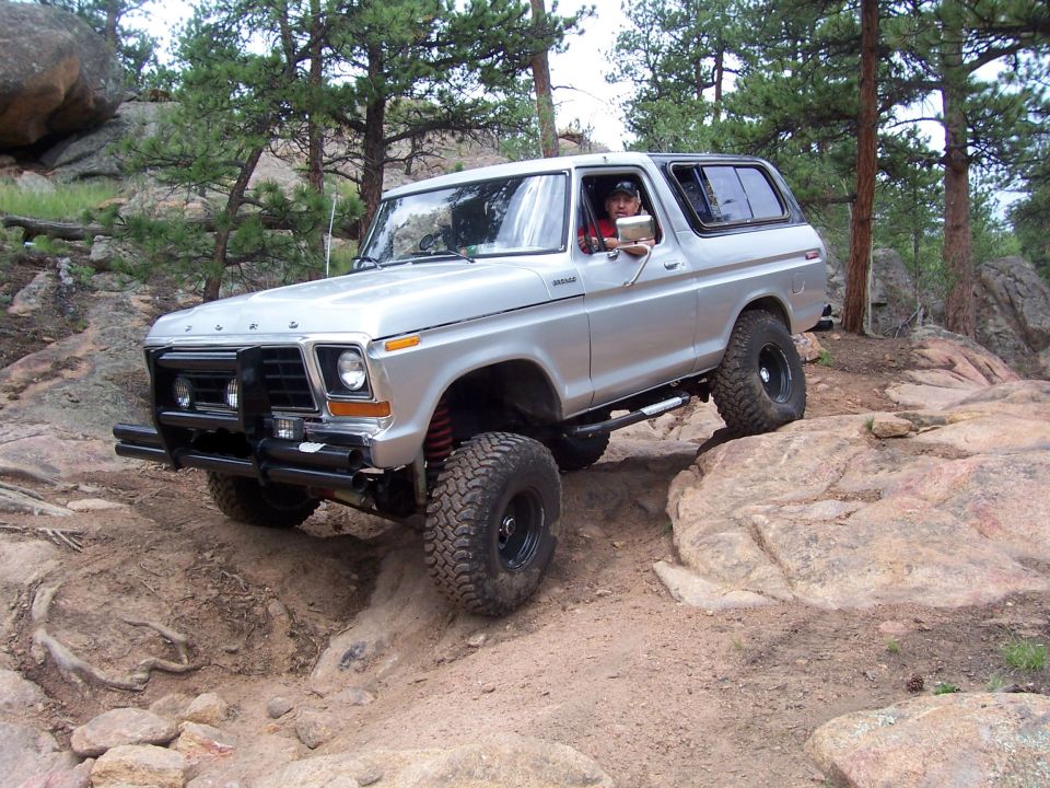 Lifted silver Ford Bronco with big mud tires climbing rocks on a forest trail