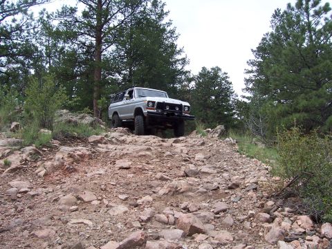 1978 Ford Bronco - 1978 Ford Bronco navigating a challenging off-road course in a wooded, rocky environment.