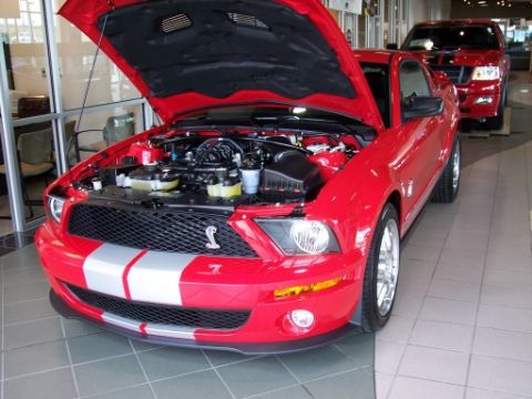 1972 Ford Ranchero - A red Ford Mustang Shelby GT500 is parked inside a showroom with its hood up, and a red Ford truck is visible in the background.