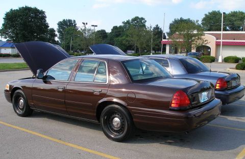 1998 Ford Crown Victoria - Two Ford Crown Victorias parked in a lot, one with its hood up.