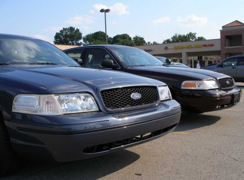 1998 Ford Crown Victoria - Two Ford Crown Victorias are parked in a lot, one dark gray and the other brown, with a building in the background.