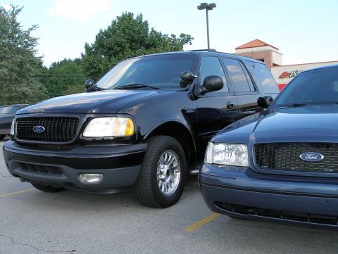 1998 Ford Crown Victoria - A blue 1998 Ford Crown Victoria police car is seen parked next to a black SUV in a parking lot.
