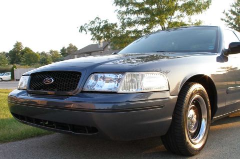 1998 Ford Crown Victoria - A dark gray Ford Crown Victoria is parked outside on a sunny day, with a house and trees in the background.