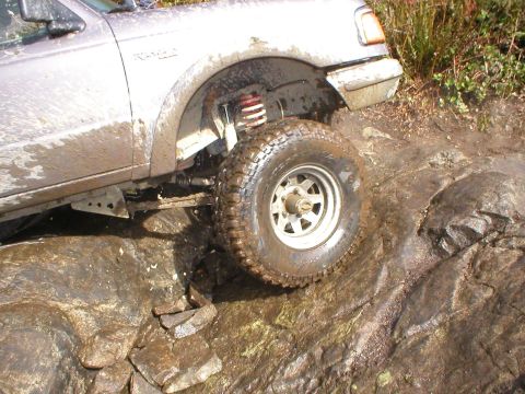 1978 Ford Bronco - A silver pickup truck navigates a rocky, muddy terrain with water splashing.