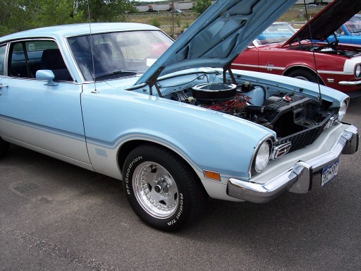 Light blue and white 1975 Ford Maverick with hood open, showing engine. Red car behind it also has hood open