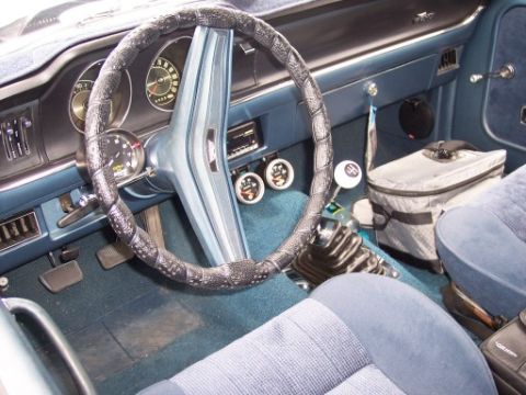 1972 Ford Ranchero - The interior of a classic car features a blue dashboard, steering wheel, and seats, with a gear shift and a cooler bag.