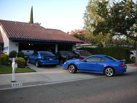 1998 Ford Crown Victoria - blue Ford Mustang coupe parked in a driveway in front of a tile-roofed house, with additional cars in the open garage behind.