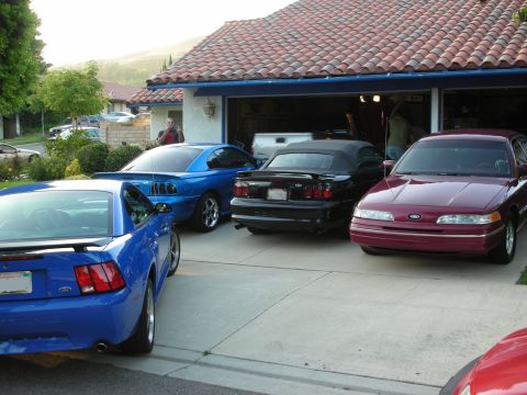 1998 Ford Crown Victoria - blue Mustang, a black convertible and a maroon Ford sedan parked in a driveway before an open garage at dusk.