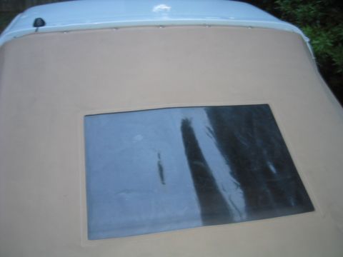 1996 Ford Bronco - Overhead close-up of the tan vinyl soft top's rear panel and its large rectangular clear vinyl window, which reflects surrounding trees. Th…