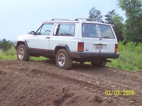 1995 Jeep Cherokee - A white Jeep Cherokee is positioned on a dirt embankment, with trees and foliage visible in the distance.