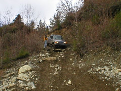 1978 Ford Bronco - A silver pickup truck navigates a challenging, off-road trail with dense vegetation on the slopes.