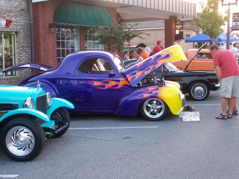 1973 Ford Ranchero - Purple 1940s Willys-style hot rod coupe with yellow-and-orange flame paint, hood tilted open to show a chrome-dressed V8, parked in profile…