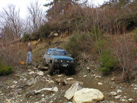 1978 Ford Bronco - A blue SUV is seen off-roading on a steep, rocky incline surrounded by trees and brush.
