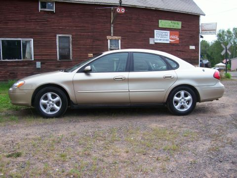 2000 Ford Taurus - A gold 2000 Ford Taurus sedan is parked on a gravel lot in front of a red wooden building with signs.