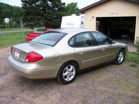 2000 Ford Taurus - A gold 2000 Ford Taurus sedan is parked in front of a garage, with a red sports car behind it.