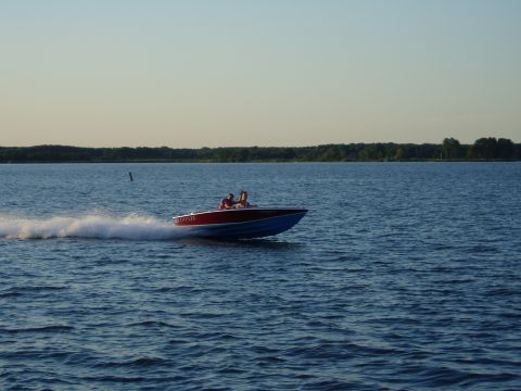 1978 Ford F-250 - A red and blue speed boat cruises across a calm lake under a clear sky.