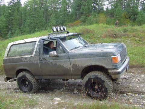 1988 Ford Bronco - Driver-side profile of a lifted green and white full-size Ford Bronco caked in dried mud, parked on a rocky wet trail with a spruce forest…