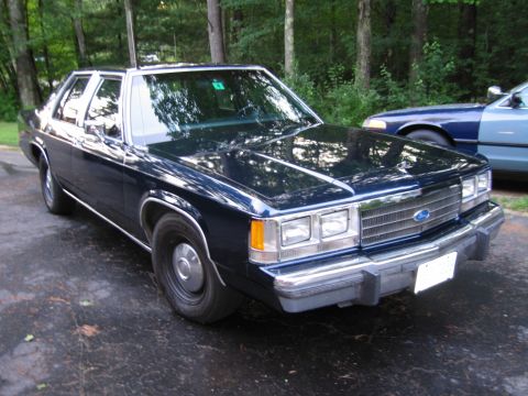 1991 Ford LTD Crown Victoria - dark blue 1980s Ford full-size sedan with a Ford grille emblem sits on a wet paved lot beside another blue car at the edge of the woods.