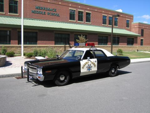 1991 Ford LTD Crown Victoria - black-and-white 1970s Dodge sedan marked HIGHWAY PATROL with a roof lightbar sits at the curb in front of Merrimack Middle School.