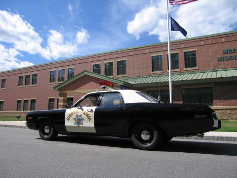 1991 Ford LTD Crown Victoria - Rear three-quarter view of a black-and-white HIGHWAY PATROL Dodge sedan parked at the curb by a brick school with flags flying overhead.