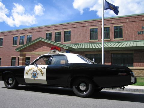 1991 Ford LTD Crown Victoria - Side profile of a black-and-white HIGHWAY PATROL Dodge sedan with a roof lightbar parked in front of a brick building under a blue sky.