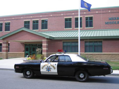 1991 Ford LTD Crown Victoria - Side view of a black-and-white HIGHWAY PATROL Dodge sedan parked by a brick school building with a flagpole and landscaping.