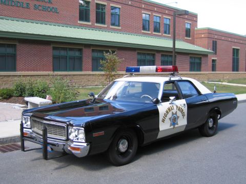1991 Ford LTD Crown Victoria - Front three-quarter view of a black-and-white HIGHWAY PATROL Dodge sedan fitted with a chrome push bar, parked beside a brick school.