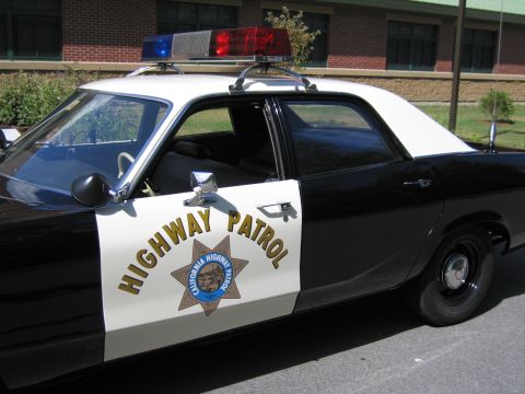 1991 Ford LTD Crown Victoria - Side view of a black-and-white CHP sedan with a front door open, displaying the HIGHWAY PATROL door decal and roof lightbar near a brick bu…