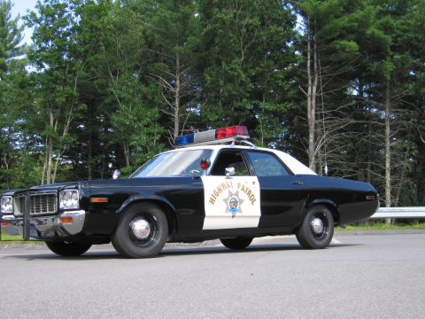 1991 Ford LTD Crown Victoria - black-and-white Highway Patrol sedan with door star emblem, lightbar and antenna, photographed front-side near woods.