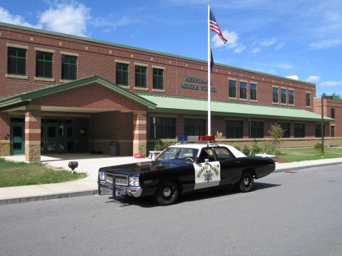 1991 Ford LTD Crown Victoria - black-and-white Highway Patrol cruiser parked at a curb in front of a brick middle school building with a flagpole.