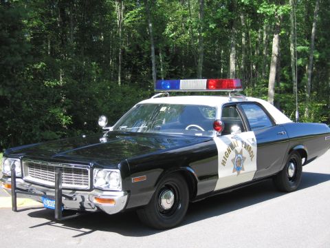 1991 Ford LTD Crown Victoria - 1970s black-and-white Highway Patrol police sedan with roof lightbar and push bar, parked on pavement near woods.