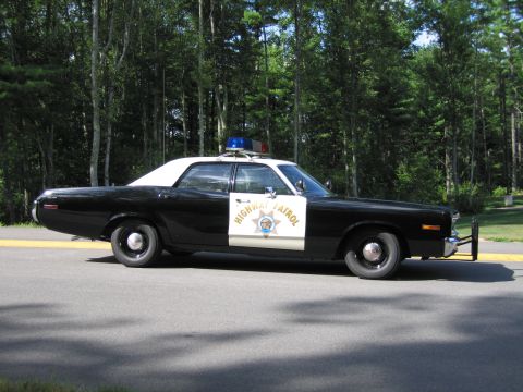 1991 Ford LTD Crown Victoria - Side profile of a black-and-white Highway Patrol police car with roof lightbar and star door emblem, parked near woods.